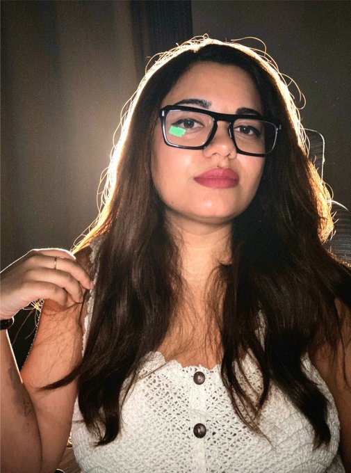 A close-up selfie of a young woman with long dark hair, wearing black-rimmed glasses and a white textured top, with a green light reflection visible in one lens.