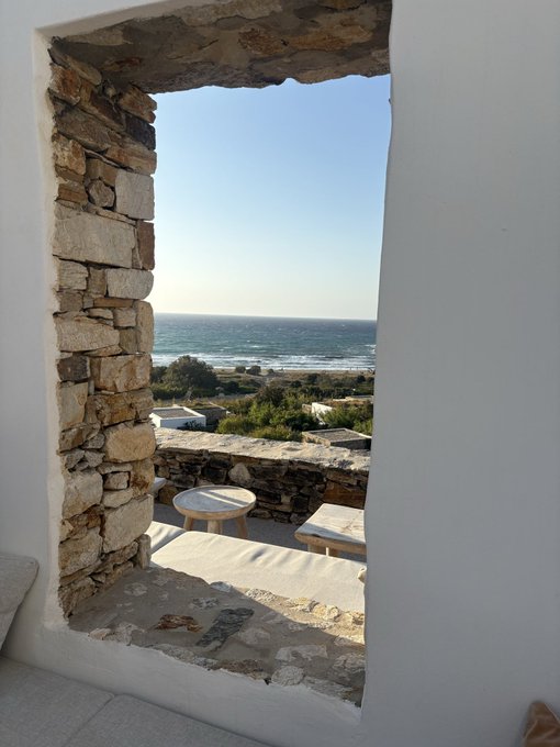 View through a stone-framed window showing a coastal scene with the ocean, beach, and outdoor seating with stone stools and tables on a terrace.