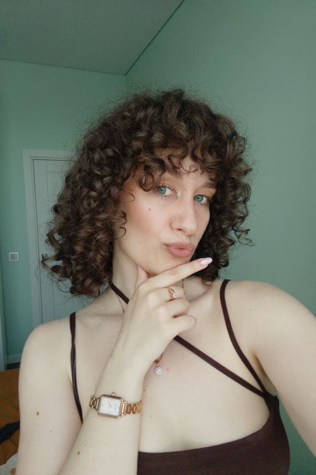 A young woman with curly brown hair poses thoughtfully with her hand on her chin, wearing a burgundy top and gold watch against a soft green wall. The contemplative pose mirrors the uncertainty of the post's question about whether Robinhood's NFT launch signals a crypto revival or precedes further market decline.