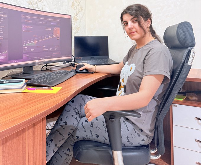 A young woman with dark hair sitting in an office chair at a curved desk with a curved monitor and laptop, turning to look at the camera.