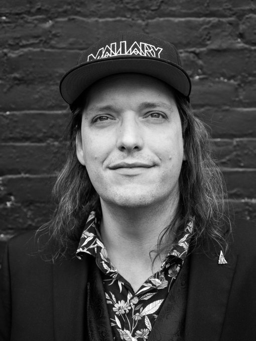 A black and white portrait photograph of a smiling man wearing a 'NUCLEAR' cap and a floral patterned shirt under a dark jacket, standing against a brick wall.