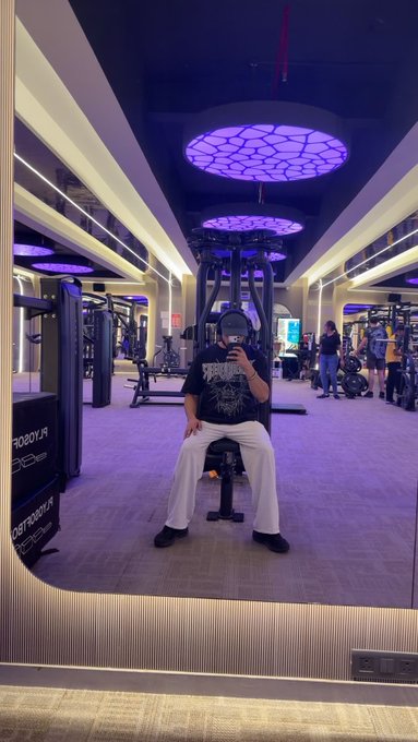 A mirror selfie taken in a gym with purple lighting and hexagonal ceiling lights, showing a person wearing a black t-shirt and white pants.