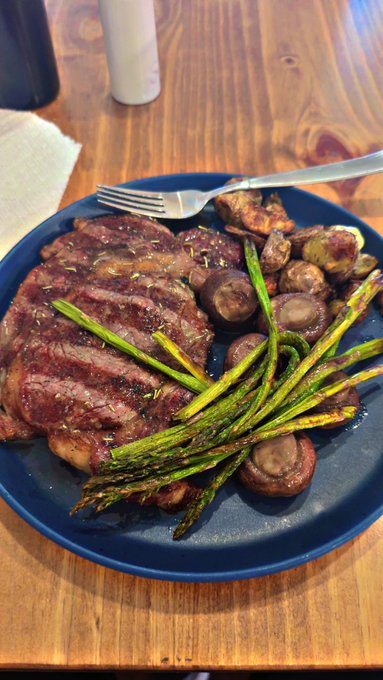 A blue plate on a wooden table holds a grilled ribeye steak garnished with rosemary, accompanied by roasted asparagus spears, sautéed mushrooms, and what appears to be roasted potatoes, with a fork resting on the rim.