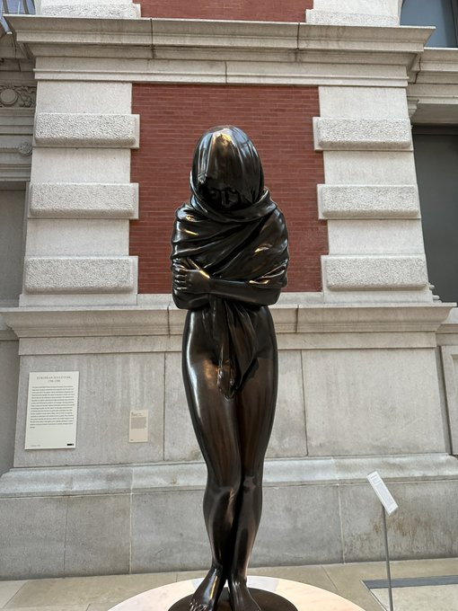 A bronze sculpture of a hooded, veiled figure stands before a stone and brick building facade, displayed outside what appears to be a museum or gallery. The accompanying plaque identifies it as part of an exhibition, fitting the post's theme of appreciating traditional, non-digital artwork.