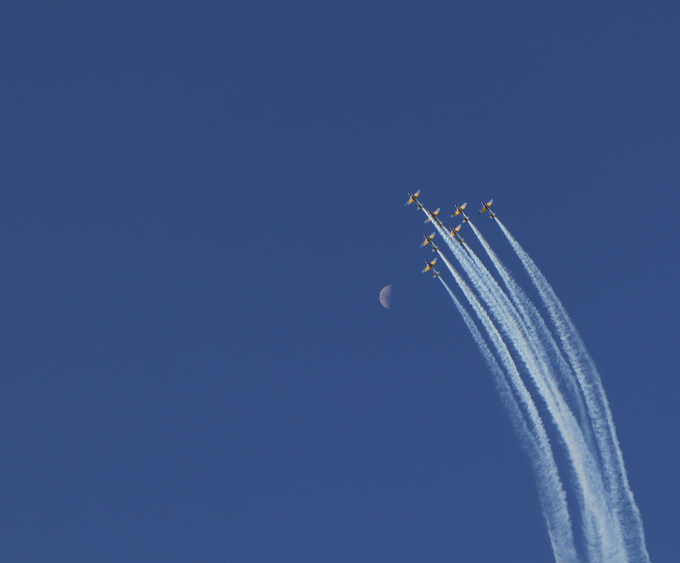 Seven jets flying in formation with white smoke trails across a deep blue sky, passing near a visible crescent moon.