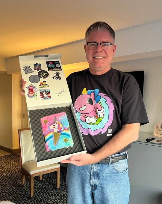 A smiling man wearing glasses and a colorful cartoon t-shirt holds an open collector's case displaying a pink unicorn cake artwork, with the interior lid covered in various pop-culture and crypto stickers including "Underpants," "MAGA," and "We the People." The image, posted alongside a caption about trust being something "you can't buy or fake" in relation to the $UFD crypto project, visually reinforces the message by showing a unique, personal collectible that couldn't be replicated or purchased through traditional means.