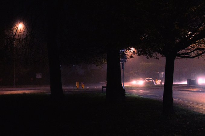 A car with headlights on drives through a misty intersection at night, illuminated by an orange streetlamp with a "30" speed limit sign visible on the left. The scene captures a darker, foggier evening at the same location compared to the original image.