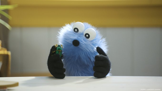 The image features a blue stuffed animal, possibly a monster or a puppet, sitting at a table with its arms outstretched as if giving a thumbs up gesture. The background of the image shows a yellow wall, which contrasts nicely with the blue color of the stuffed animal and adds visual interest to the scene. There is also an object on the table that appears to be a green toy or a small figurine.