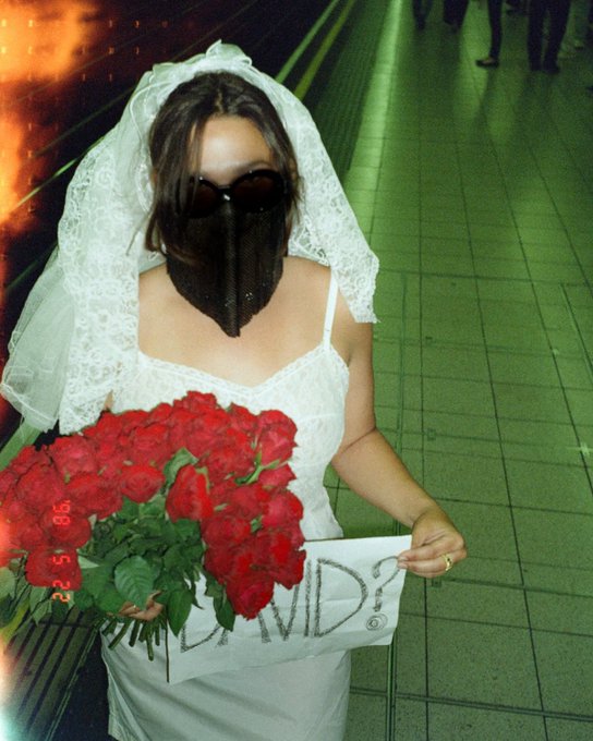 A person in a white lace wedding dress and veil wears a black plague doctor mask, holds a bouquet of red roses and a sign reading 'COVID?', with a timestamp '9.5.20'.