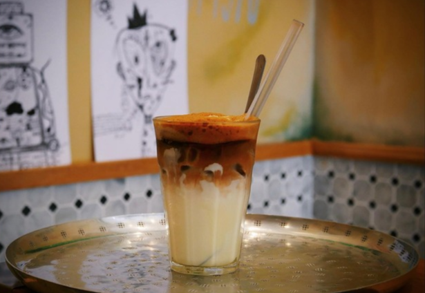 A tall glass of layered iced coffee with a thick frothy top and distinct milk and coffee sections sits on a silver tray with straws and a spoon, set against a yellow wall decorated with black-and-white sketches.