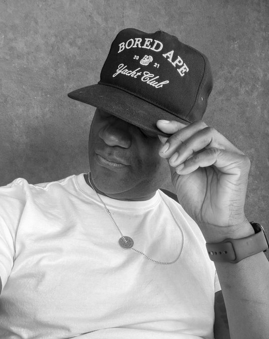 A black-and-white photograph of a man wearing a white t-shirt, a silver chain necklace with a pendant, a smartwatch, and a dark baseball cap that reads 'BORED APE 20 21 Yacht Club', with the brim pulled down to obscure his eyes.