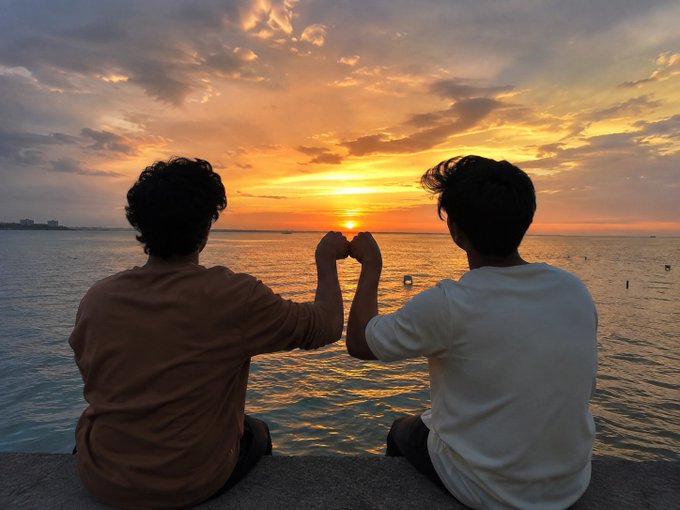 Two friends sit side by side at the water's edge, forming a heart shape with their hands against a golden sunset, symbolizing the silent bond and unwavering trust shared between best friends.
