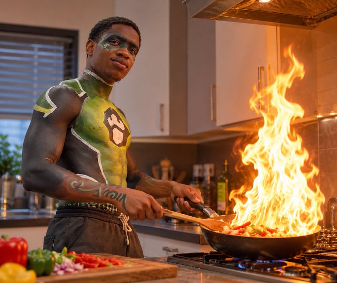 A young man wearing green and black body paint resembling a superhero costume is cooking vegetables in a flaming wok in a kitchen.