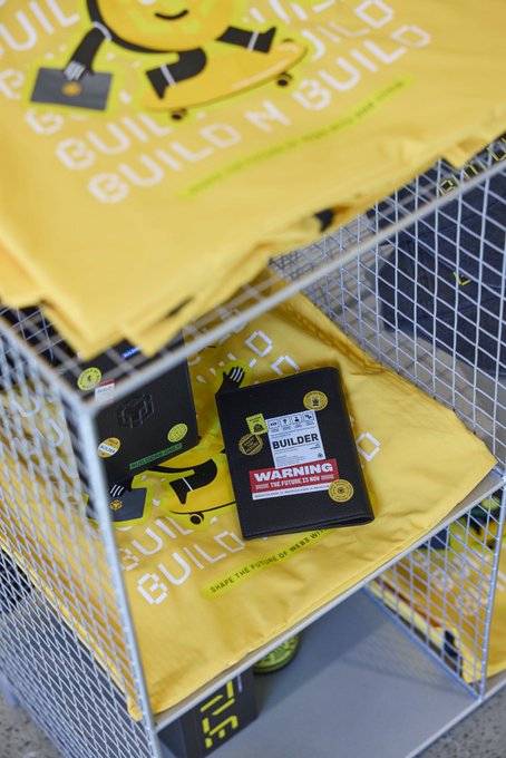 Yellow $BUILDER merchandise featuring the BNB Chain mascot is displayed in a wire basket, including t-shirts with "BUILD N BUILD" text and merchandise labeled with "BUILDER" and a warning sticker reading "WARNING MAY GET BULLISH AS HELL."