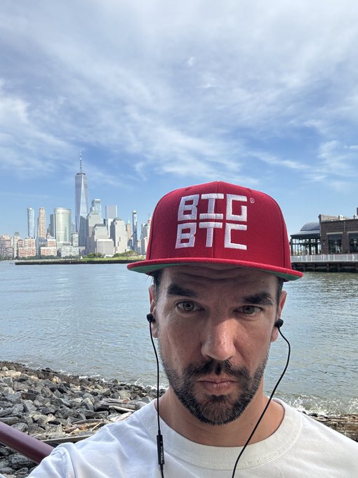 A man wearing a red BTC BTC cap and earphones takes a selfie in front of the New York City skyline with One World Trade Center visible across the water.