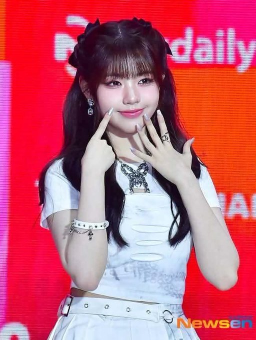 A young woman with long dark hair and bangs poses playfully, pointing at her cheeks while wearing a white crop top, statement necklace, and beaded bracelet; 'Newsen' watermark visible.
