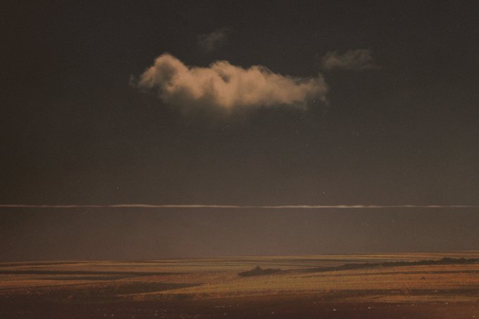 A night landscape photograph showing a dark, expansive field or beach under a vast sky, highlighted by a single, warmly lit cloud floating in the center.
