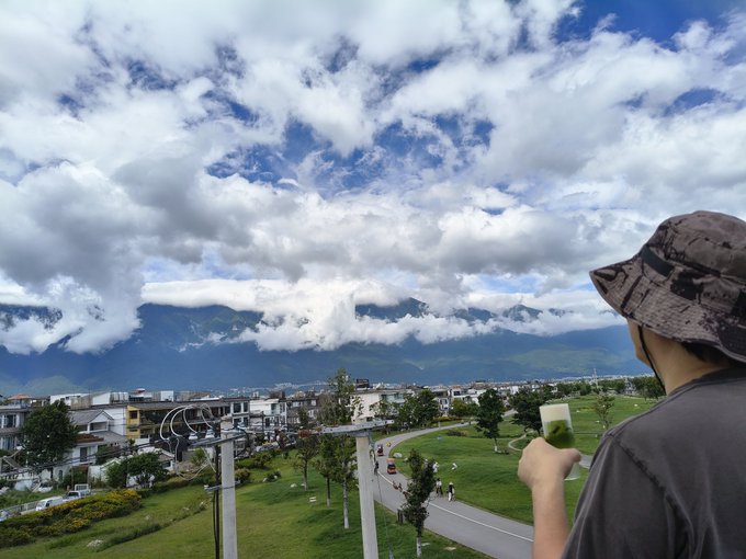 A person in a bucket hat holds up a phone to capture the sweeping mountain landscape and town below, beneath a dramatic sky of clouds and blue. The scene mirrors the "good morning" sentiment, capturing a peaceful sunrise moment that contrasts with the hopeful buzz of crypto dreams.