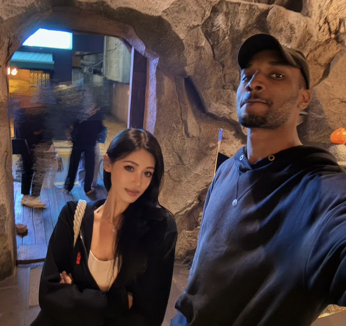 A young woman with long dark hair, wearing a black cardigan and white top, stands next to a tall man in a navy hoodie and black cap, posing for a selfie inside a cave-like structure with rocky walls. Behind them, other patrons can be seen in the dimly lit interior space.