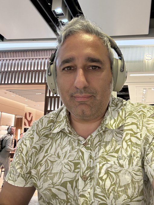 A man wearing a green and white tropical-print shirt and light green over-ear headphones takes a selfie indoors.