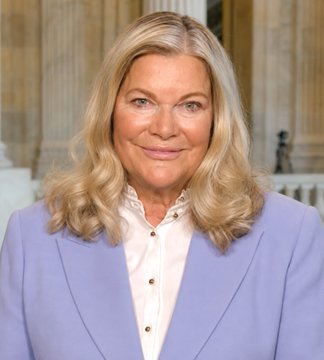 A portrait photograph of a blonde woman wearing a light purple blazer over a pink ruffled blouse, smiling slightly in front of a classical columned background.