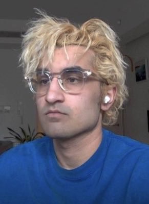 A young man with curly bleached blonde hair, clear-framed glasses, and a white earbud is shown in a close-up selfie wearing a blue shirt. The image appears to depict FrankDeGods, referenced in the breaking news about a leaked brief revealing influencers were targeted in a fraudulent marketing campaign involving fake defamatory images against creators.