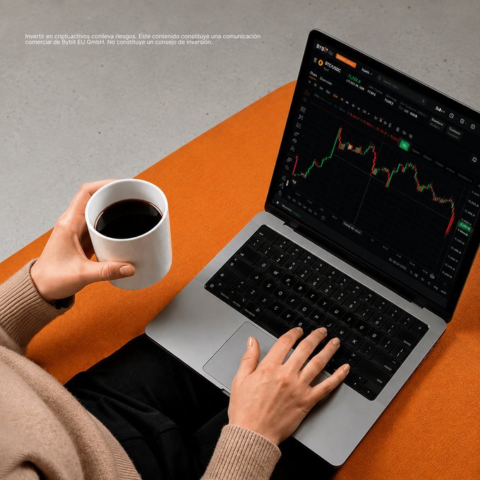 A photo of someone holding a white coffee mug while using a laptop displaying Bybit exchange with BTC/USDC trading chart, with Spanish disclaimer text about crypto investment risks from Bybit EU GmbH.