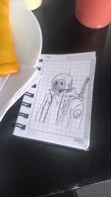 A pencil sketch in a small spiral notebook depicts a robotic character with an antenna and a sword on its back, sitting beside a plate of food and a red mug on a dark surface.