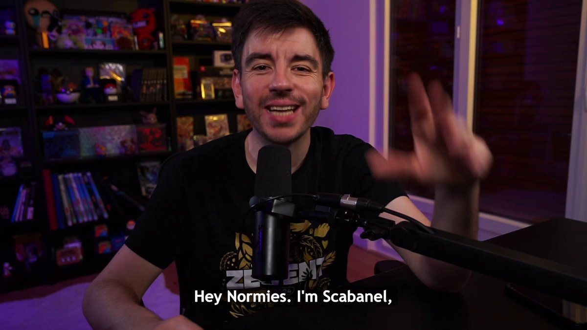 The image shows a man sitting at a desk holding a microphone while wearing a black t-shirt with yellow text that says "hey normies, i'm scabane,". The background of the image features a bookshelf filled with various items such as books and DVDs. There is also a caption in the image that reads "During the @NFC summit, I showed the tool I built for Normies Holders and people were pretty impressed (and I was flattered!) Today, Normies Nexus evolved a lot with a lot of new features for both holders but also curious ones!".