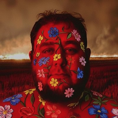 A surreal portrait of a man's face covered in colorful floral tattoos against a red background with a smoky, fiery landscape.