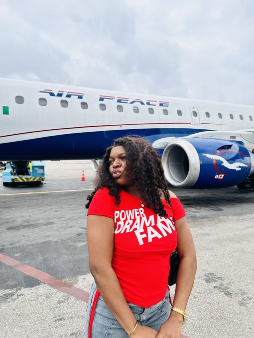 A woman wearing a red 'POWER DREAM FAN' t-shirt stands on a tarmac in front of an airplane with 'PEACE' written on its fuselage.