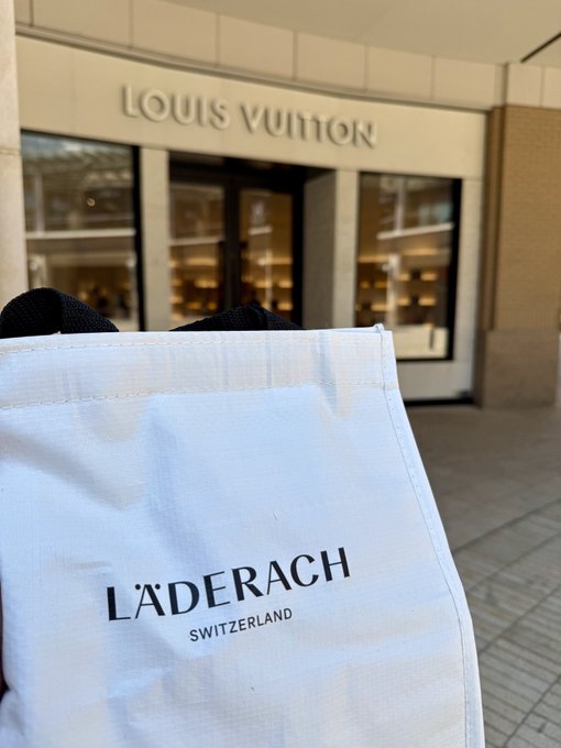 A first-person perspective photo showing a hand holding a white shopping bag with the 'LÄDERACH SWITZERLAND' logo in front of a 'LOUIS VUITTON' storefront.