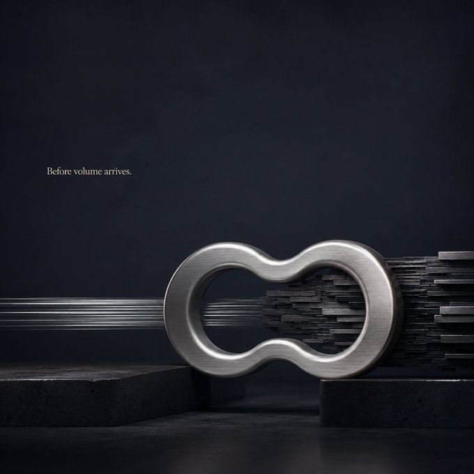 A sleek metallic figure-8 shaped object displayed on dark stone pedestals against a black background, with the text 'Before volume arrives.' on the left side.