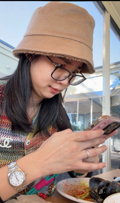 A woman wearing a tan fuzzy bucket hat, glasses, and a colorful knit top with a Chanel logo is looking down at her phone while eating a seafood dish at a restaurant. The image accompanies a post greeting MoonPay but shows no direct relation to the company beyond the casual dining scene.