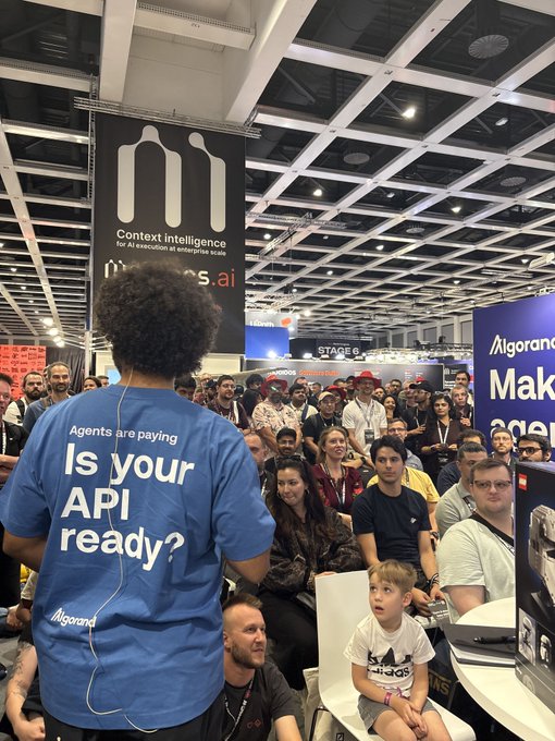 A scene from a busy tech convention shows a person in a blue shirt reading 'Agents are paying - Is your API ready? Algorand' addressing a seated crowd, with 'mess.ai' and 'STAGE 6' banners visible in the background.