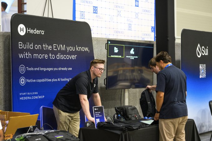 Two men work at a Hedera booth at a conference, with signage reading 'Build on the EVM you know, with more to discover' and a neighboring Sui banner.