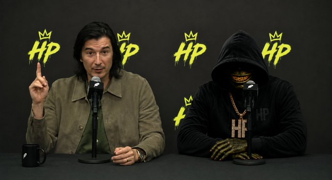 Two men sit at a press conference table with microphones in front of a black backdrop displaying yellow 'HP' logos with crowns; the man on the right wears a black hoodie with his face covered by a gold skull mask and matching gold gloves.