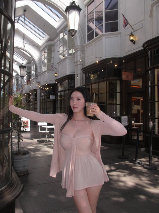A woman in a pink outfit holds an iced coffee drink in a bright, elegant indoor passageway featuring glass ceilings and string lights.