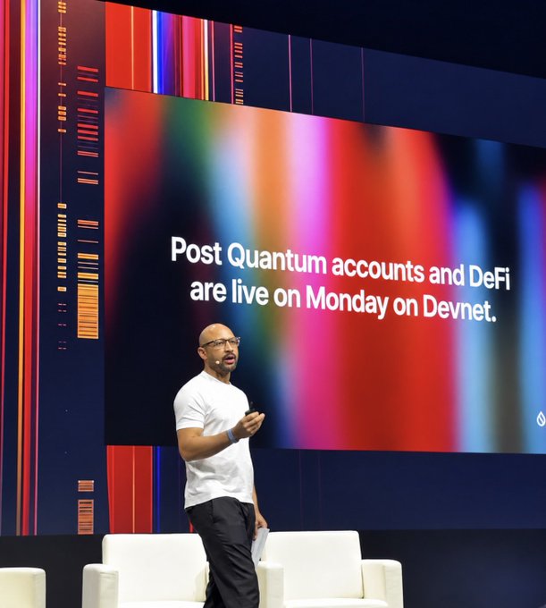 A bald man with glasses and a beard, wearing a white t-shirt, speaks on a stage in front of a large screen displaying the text 'Post Quantum accounts and DeFi are live on Monday on Devnet.'
