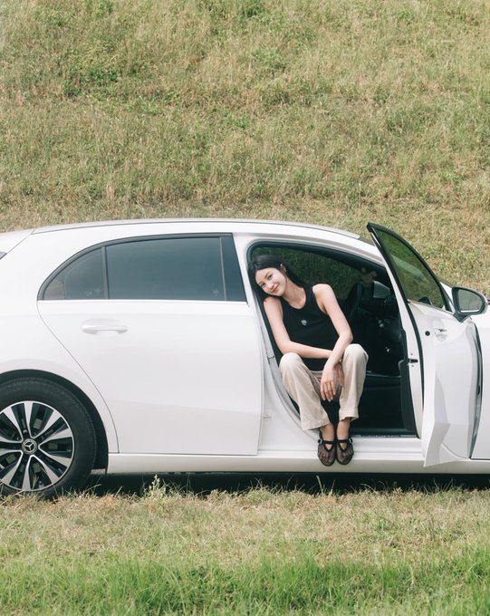 A woman sits in the open doorway of a white sedan parked on a grassy area, smiling at the camera.