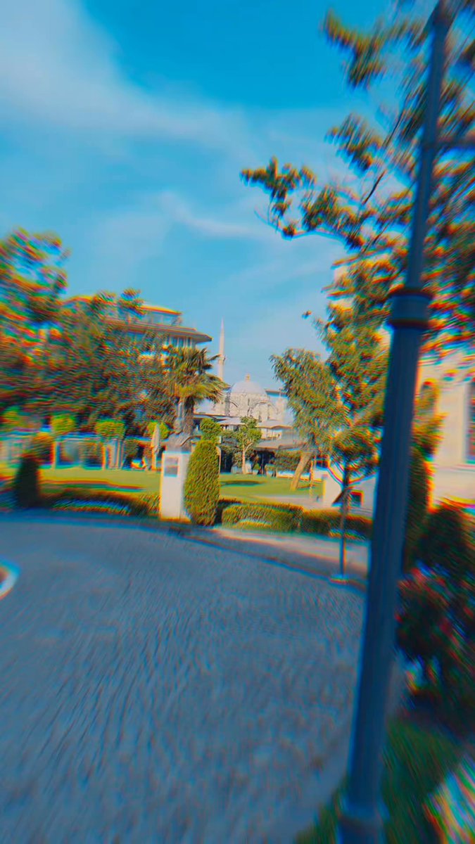 The image shows an outdoor scene with a cobblestone street, trees, and buildings in the background. A black lamppost stands near the center of the frame, while a white building can be seen further back. The sky above appears hazy, creating a soft and dreamy atmosphere. In the foreground, there is a bench where one could sit to enjoy the view or take a break from walking.