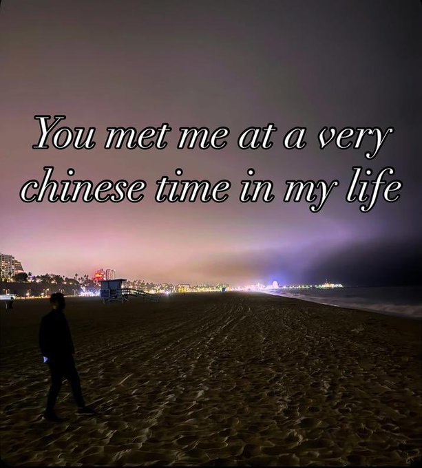 A night beach scene with a lone figure walking on the sand and lit-up buildings along the shoreline, overlaid with the text "You met me at a very chinese time in my life".