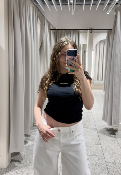 Mirror selfie of a young woman with long wavy hair wearing a black sleeveless crop top and white jeans in a fitting room with grey curtains.