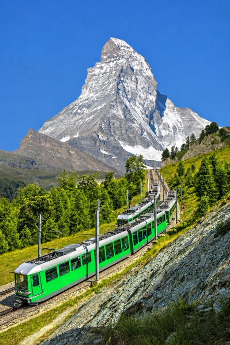 A bright green passenger train winds through a grassy alpine mountainside toward the snow-capped Matterhorn peak under a clear blue sky.