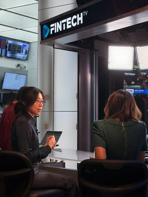 An on-camera interview takes place in a FinTech TV studio at the NYSE, featuring a woman with glasses seated across from another interviewee, with the "FINTECH TV" logo prominently displayed above. The scene illustrates the post's context of OP Labs' CEO discussing centralized exchange infrastructure on the NYSE floor.