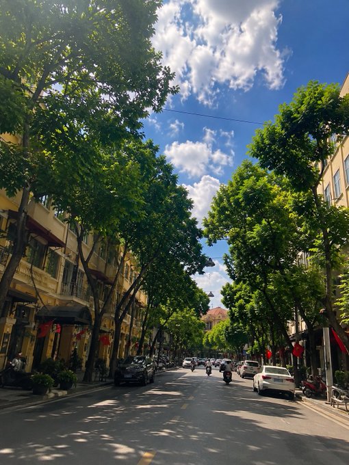 A sunlit tree-lined street in Vietnam, likely Hanoi, with parked cars and motorbikes under a bright blue sky dotted with clouds, capturing the hot, pleasant weekend weather mentioned in the post. No legible text or signage is visible in the image itself.