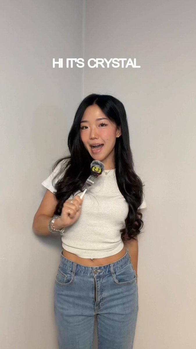 A young woman with long black hair wearing a white top holds a silver spoon, which has an electronic device attached to it that plays music when she says "hi its crystal." The background appears to be a gray wall adorned with a painting of a bird. This setting adds an artistic element to the image and reinforces the theme of the post about cryptocurrency news and trading.