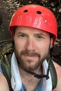 A close-up selfie of a bearded man wearing a red safety helmet and white neck fan, appearing to be at an outdoor activity location.