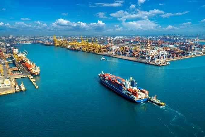 An aerial view shows a large container ship being guided by a tugboat through a busy port with extensive container terminals and yellow gantry cranes under a blue sky.