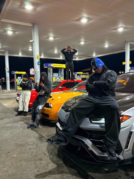 A group of young people posing around a yellow and a silver Lamborghini at a gas station at night, with one person sitting on the silver car's hood and another standing on a gas pump canopy.